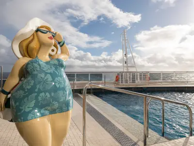 Eine farbenfrohe Skulptur einer Frau in Badekleidung, Sonnenhut und Sonnenbrille steht am South Beach Pool an Bord der MSC Seaside. Im Hintergrund sind das glitzernde Meer und ein teilweise bewölkter Himmel zu sehen.