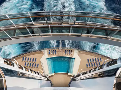 Vogelperspektive auf das offene Deck der MSC Seaside mit einem großen blauen Pool und vielen Liegestühlen. Eine gläserne Brücke spannt sich über das Deck, darunter schäumt das Meer. Wenige Passagiere entspannen auf den Liegen.