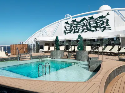 Ein sonniger Poolbereich auf dem Oberdeck der MSC Seaside. Türkisfarbenes Wasser, helle Sonnenliegen und beeindruckende Skulpturen von Delfinen und Seepferdchen schmücken die Szene. Im Hintergrund ist das Meer sichtbar.