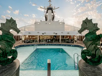 Blick auf den Poolbereich des MSC Yacht Club auf der MSC Seaside. Zwei grüne Seepferdchen-Skulpturen flankieren den rechteckigen Pool. Im Hintergrund befindet sich eine moderne Bar mit Sitzgelegenheiten unter einem teilweise überdachten Bereich.