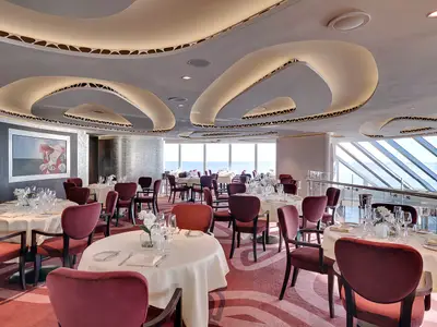 Elegantes Restaurant an Bord der MSC Seaside. Gedeckte Tische mit weinroten Stühlen und Blick auf das Meer. Moderne Decke mit geschwungenen Lichtinstallationen, Kunstwerke an den Wänden.