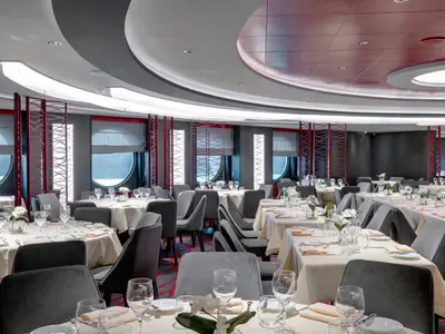 Innenansicht des Seashore Restaurants an Bord der MSC Seaside, das eine Reihe von elegant gedeckten Tischen mit grauen Stühlen und roten Zierelementen zeigt. Durch die runden Fenster ist das Meer zu sehen.