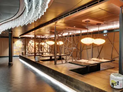 Blick in das asiatische Spezialitätenrestaurant "Asian Market Kitchen" an Bord der MSC Seaside. Es zeigt Tische in Kabinen mit Holzlatten-Trennwänden und großen ovalen Pendelleuchten. Ein langer Chandelier schmückt den Gang.
