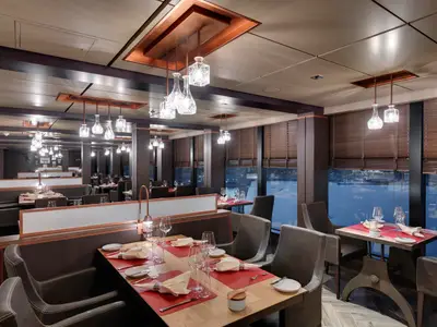 Blick in ein stilvolles Spezialitätenrestaurant an Bord der MSC Seaside bei Abendstimmung, mit fertig gedeckten Tischen und grossen Fenstern, die eine Hafenansicht zeigen.