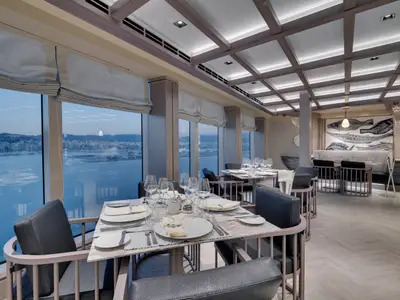 Blick ins Ocean Cay Restaurant der MSC Seaside. Elegante Tische am Fenster mit Hafenblick. Eine Wand zeigt Fisch-Mosaik. Moderne Beleuchtung.