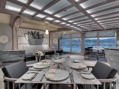 Das Ocean Cay Spezialitätenrestaurant an Bord der MSC Seaside zeigt elegant gedeckte Tische, eine Bar und grosse Fenster mit Blick auf eine belebte Hafenstadt bei Dämmerung.