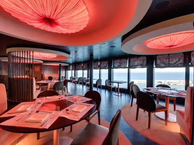 Modernes Spezialitätenrestaurant an Bord der MSC Seaside mit gedeckten Tischen, bequemen Sitzen und roten Deckenleuchten. Grosse Fenster bieten Meer- und Hafenblick.