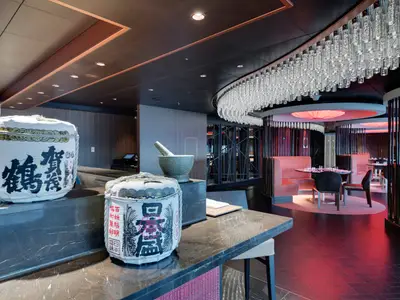 Restaurant an Bord der MSC Seaside. Zwei Sake-Fässer auf einer Theke. Elegante pinke Sitzbereiche mit Kristallleuchter und roter Beleuchtung.