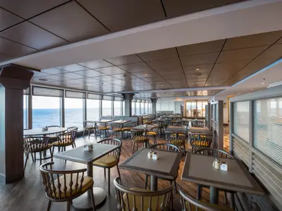 Ein heller Restaurantbereich an Bord der MSC Seaside mit zahlreichen Tischen und Stühlen. Große Fenster bieten einen Panoramablick auf das weite, blaue Meer und den Horizont. Die Einrichtung ist modern und einladend.