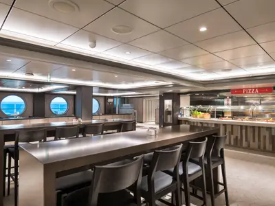 Das Marketplace Buffetrestaurant der MSC Seaside. Lange Tische, runde Bullaugen mit Meerblick und eine Pizzastation sind zu sehen.
