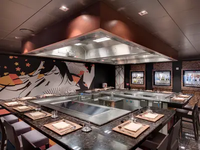 Modernes Teppanyaki-Restaurant an Bord der MSC Seaside. Glänzende Kochstationen, dunkle Tische mit Gedeck, asiatisches Wandbild, beleuchtete Getränkekühlschränke prägen den Raum.