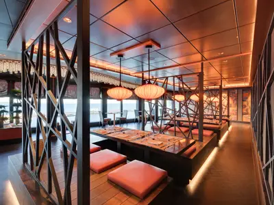 Innenansicht des stilvollen Asian Market Kitchen Spezialitätenrestaurants an Bord der MSC Seaside. Elegante Holztische mit roten Sitzkissen, kunstvolle Laternen und Panoramasicht auf das Meer.