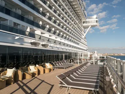 Großes Sonnendeck der MSC Seaside mit vielen braunen Liegestühlen und Rattansesseln auf Holzboden, flankiert von Glasfronten und unzähligen Balkonkabinen. Im Hintergrund ist eine weitläufige Küstenstadt am Meer unter blauem Himmel zu sehen.