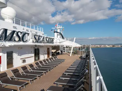 Das geräumige Sonnendeck der MSC Seaside ist mit zahlreichen braunen Liegestühlen bestückt. Im Hintergrund ist das Meer und eine Küstenstadt zu sehen.