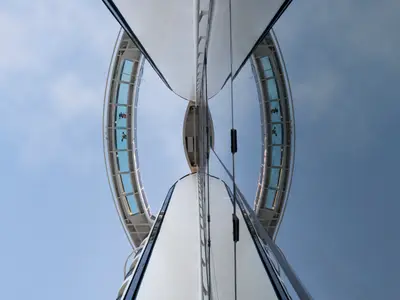 Blick von unten auf die gläserne Bridge of Sighs der MSC Seaside. Personen laufen auf dem Steg. Eine Spiegelung erzeugt eine symmetrische Brückenansicht vor blauem Himmel.