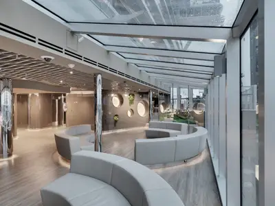 Moderne Lounge im MSC Aurea Spa an Bord der MSC Seaside mit hellgrauen, geschwungenen Sofas, glänzenden Säulen, einer Wanddekoration mit runden Nischen und Pflanzen sowie einem großen Glasdach.