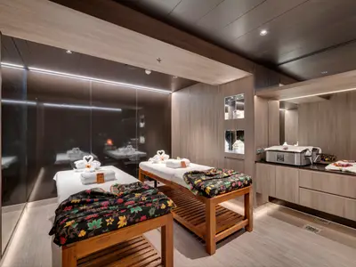 Ein Spa-Behandlungsraum an Bord der MSC Seaside mit zwei Massageliegen, dekoriert mit Handtuch-Schwänen und floralen Decken. Holzwände, Spiegel und indirekte Beleuchtung schaffen eine ruhige Atmosphäre. Ein Hot-Stone-Wärmer steht bereit.