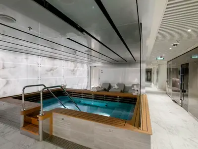 Das luxuriöse Spa der MSC Seaside zeigt einen großen, beleuchteten Pool mit Holzdeck, umgeben von eleganten Liegen und Wänden aus hellem Marmor. Ein heller Gang führt zu weiteren Behandlungsräumen.