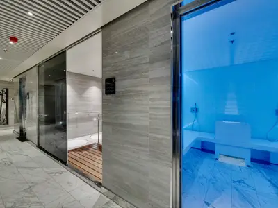 Ein heller, moderner Wellnessbereich an Bord der MSC Seaside. Links sind Duschkabinen mit Holzboden zu sehen, rechts ein blau beleuchteter Raum mit Sitzbänken und Duscharmaturen an den Wänden, vermutlich ein Dampfbad oder Eisraum. Die Wände sind mit hellen Marmorfliesen verkleidet, der Boden mit weissem Marmor. Das Licht ist überwiegend kühles Blau und Weiss.