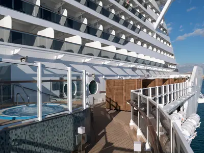 Ein blauer Whirlpool auf dem Holzdeck der MSC Seaside, umgeben von Glasgeländern. Darüber sind die Balkonkabinen sichtbar. Im Hintergrund das weite Meer und eine Küstenlinie unter blauem Himmel.