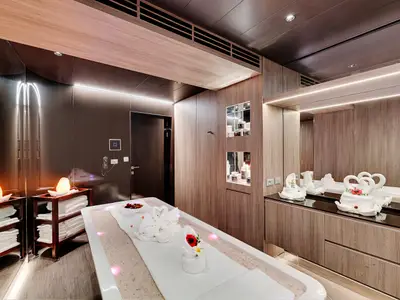 Ein luxuriöser Spa-Behandlungsraum an Bord der MSC Seaside. Im Vordergrund ein Behandlungsbett mit Handtuch-Schwänen und Blumen, umgeben von warmem Licht. Eine Ablage mit weiteren weißen Handtüchern und Salzkristalllampen steht daneben. Die Wände sind mit Holzpaneelen und dunklen, reflektierenden Oberflächen verkleidet. Spiegel reflektieren Kosmetikprodukte.