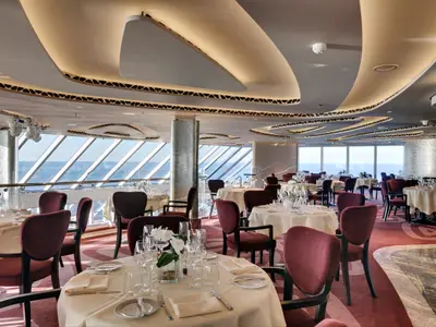 Elegantes Restaurant an Bord der MSC Seaside mit stilvoll gedeckten Tischen und bordeauxroten Stühlen. Große Panoramafenster bieten einen weiten Blick auf das offene Meer. Die moderne, geschwungene Deckenbeleuchtung und der rote Teppichboden schaffen eine luxuriöse Atmosphäre. Der Raum ist ohne Personen.