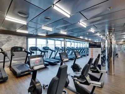Modernes Fitnessstudio an Bord der MSC Seaside mit Technogym-Laufbändern und Ergometern. Große Fensterfronten bieten Meerblick. Helle, offene Atmosphäre mit Holzboden und Wandgrafik. Ohne Personen.