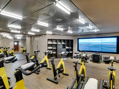Ein modernes Fitnessstudio an Bord der MSC Seaside mit zahlreichen gelb-schwarzen Spinning-Rädern. Im Hintergrund eine grosse Videowall mit einer Landschaftsaufnahme und Regalen voller Handtücher. Spiegelflächen an der linken Wand.