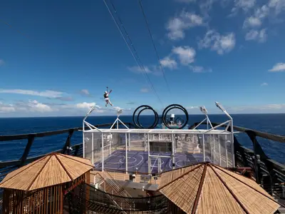 Eine Person gleitet an einer Zip-Line über dem Sportdeck der MSC Seaside mit Basketballfeld und Blick auf das weite Meer.