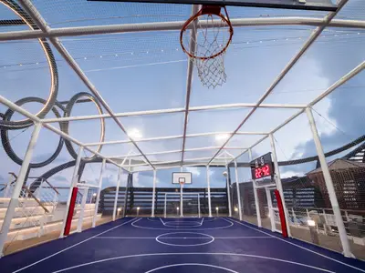 Eine leere Sportarena an Bord der MSC Seaside, umgeben von Netzen. Sie zeigt einen Basketballkorb, ein Fussballtor und eine Anzeigetafel. Die blaue Spielfläche ist für verschiedene Sportarten markiert. Im Hintergrund sind weitere Schiffstrukturen und eine auffällige Lichtinstallation zu sehen. Der Himmel ist bewölkt.