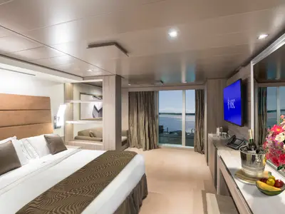Eine geräumige MSC Yacht Club Deluxe Suite an Bord der MSC Seaside mit einem komfortablen Doppelbett, einem einladenden Balkon mit Meerblick und einem Willkommenspaket bestehend aus Wein, Eis und Obst. Das moderne Design und die warme Beleuchtung schaffen eine elegante Atmosphäre.