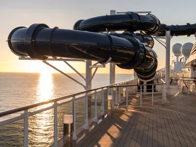 Schwarze Wasserrutschen der MSC Seaside ragen über dem Meer. Goldener Sonnenuntergang spiegelt sich auf dem Wasser. Sonnendeck mit Holzplanken und weissen Geländern.