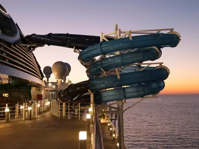 Blick auf die Wasserrutschen an Bord der MSC Seaside im warmen Licht des Sonnenuntergangs. Die blaue und schwarze Rutsche ragen über das Meer. Das beleuchtete Deck ist menschenleer.