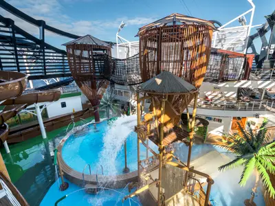 Der Aquaventure Park auf der MSC Seaside. Wasserrutschen, ein Turm mit Kippeimer und Kletternetze sind zu sehen. Wasser spritzt in den Pool. Im Hintergrund das Meer und Sonnendecks.
