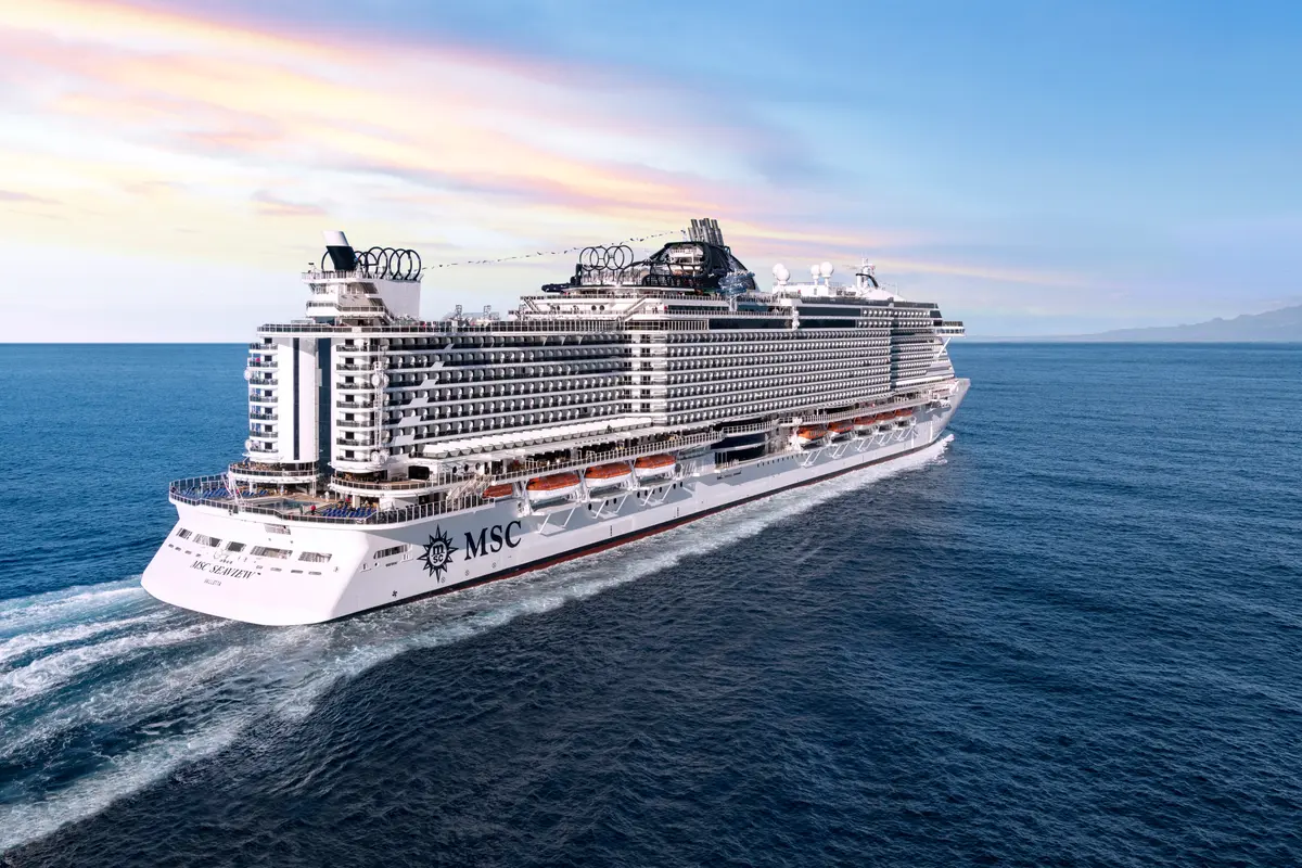 Luftaufnahme des megagroßen Kreuzfahrtschiffes MSC Seaview, das auf offener See fährt und weiße Schaumspuren im blauen Wasser hinterlässt. Im Hintergrund sind Berge und ein farbiger Himmel zu sehen.