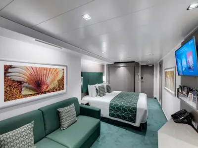 Blick in eine helle Balkonkabine an Bord der MSC Seaview mit großem Doppelbett, dunkelgrünem Kopfteil, passendem Sofa und einem Wandbild mit Muschelmotiv.