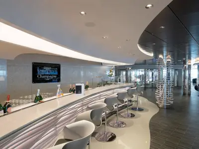 Eine moderne und elegante Champagne Bar an Bord der MSC Seaview. Die geschwungene Bar Theke ist mit eisgekühlten Champagnerflaschen und Gläsern bestückt. Dahinter ein beleuchtetes "Champagne Bar" Schild und stylische Barhocker. Das Interieur ist von glänzenden Oberflächen und futuristischen Säulen geprägt.