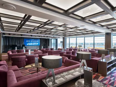 Geräumige Seaside Lounge an Bord der MSC Seaview mit zahlreichen violetten Samtsofas, einem Flügel auf einer Bühne und großen Fenstern mit Meerblick. Die Decke ist mit Lichtpaneelen versehen, und auf Bildschirmen ist das Schiff "MSC Seaview" zu sehen.