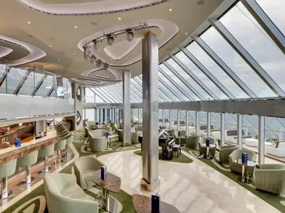 Weitwinkelaufnahme der stilvollen Top Sail Lounge an Bord der MSC Seaview mit Bar, Klavier, Sitzgelegenheiten und Panoramablick durch grosse Fenster.