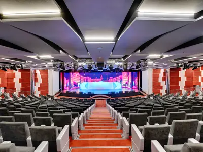 Ein Weitwinkelbild des Odeon Theaters an Bord der MSC Seaview. Die Bühne ist hell beleuchtet mit einem futuristischen Stadtbild auf der LED-Leinwand und einem hellblauen Podest davor. Zahlreiche Sitzreihen in dunklem Grau sind zu sehen, sowie rot-weiße Wandverkleidungen und eine moderne Deckenkonstruktion.