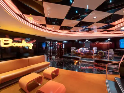 Unterhaltungsbereich der MSC Seaview mit 'Bowling'-Schild, orangen Sofas und F1-Simulator. Moderne Einrichtung mit reflektierenden Decken und Neonlicht.