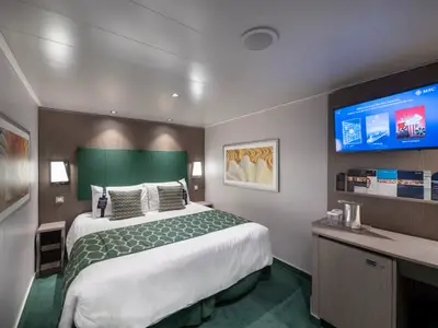 Moderne Innenkabine an Bord der MSC Seaview. Doppelbett mit grüner Tagesdecke, Nachttische mit Lampen. Gegenüber ein Flachbild-TV über Minibar. Helle Wände, Holzakzente, Kunstwerke.