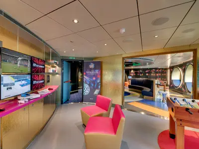 Farbenfroher Teen Club an Bord der MSC Seaview. Gaming-Ecke mit Bildschirmen und Konsolen. Loungebereich mit Foosballtisch und runden Fenstern. Goldene Wände.