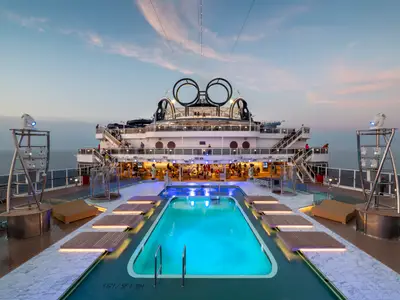 Der blau beleuchtete Panorama Pool der MSC Seaview bei Abenddämmerung. Moderne Decks, beleuchtete Bars mit Gästen im Hintergrund. Die ikonische Schiffskrone und das Meer sind sichtbar.