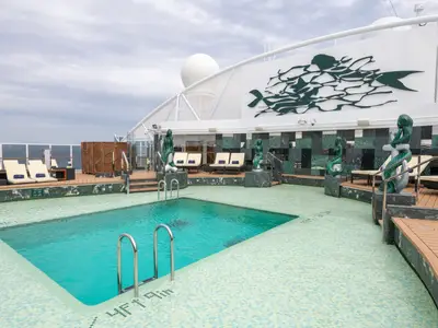 Blick auf den Poolbereich des MSC Yacht Club an Bord der MSC Seaview. Türkisblaues Wasser, umgeben von Liegestühlen und grün getönten Meerjungfrau-Skulpturen vor einer marmorierten Wand.