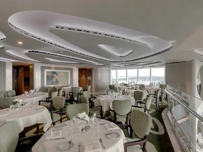 Innenansicht des eleganten MSC Yacht Club Restaurants an Bord der MSC Seaview mit weiss gedeckten Tischen, bequemen Sesseln und grossen Fenstern, die einen Blick auf einen Hafen bieten. Eine Kunstinstallation an der Decke und ein grosses Gemaelde zieren den Raum.
