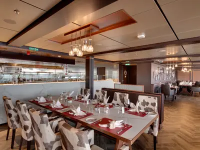 Ein elegantes Steakhouse, Butcher's Cut, an Bord der MSC Seaview. Im Vordergrund ein langer Tisch mit Kuhfell-Stühlen, im Hintergrund die offene Küche und weitere Sitzbereiche. Alles ist festlich gedeckt und gut beleuchtet.