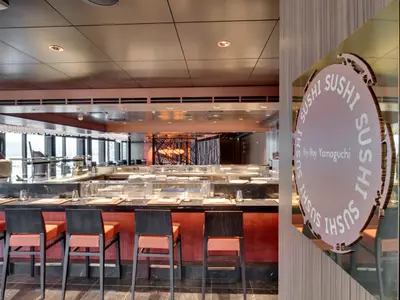 Innenansicht der Asian Market Kitchen Sushi Bar By Roy Yamaguchi an Bord der MSC Seaview mit dunkler Theke, orangefarbenen Hockern und großen Fenstern im Hintergrund.