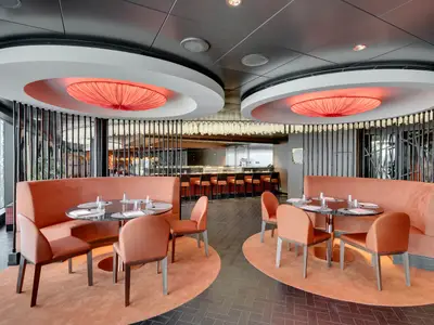 Elegantes Pan-asiatisches Spezialitätenrestaurant Asian Market Kitchen an Bord der MSC Seaview mit orangefarbenen Sitzbänken, runden Tischen und beeindruckenden Deckenlampen.