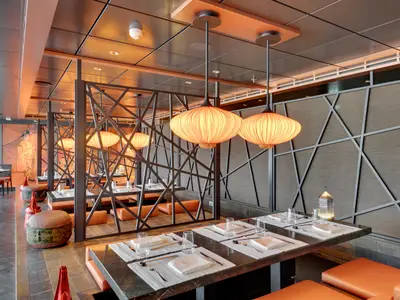 Blick in die Asian Market Kitchen an Bord der MSC Seaview. Der Speisesaal ist mit dunkelbraunen Trennwänden und orangefarbenen Deckenlampen dekoriert. Tische sind eingedeckt, an der Wand ein Gemälde mit einer Geisha.